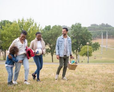 A joyful family walking through a park on a sunny day, carrying a picnic basket and a ball.